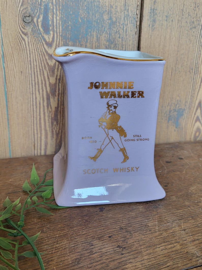 Op de afbeelding: Een paarse keramische kan met een gouden rand en een Johnnie Walker-logo. Het logo toont een man in een wandelende pose met de tekst "Born 1820 - Still Going Strong" en "Scotch Whisky".