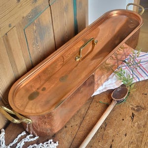 May include: A copper rectangular cooking pot with a lid and brass handles. The pot is long and narrow, with rounded ends. A wooden spoon rests nearby. The pot is on a wooden surface.