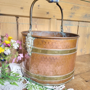 Puede incluir: Un cubo de cobre con un borde de latón y un asa decorativa. El asa está hecha de cerámica con un diseño floral azul y blanco. El cubo está sobre un mantel de encaje blanco.