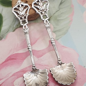 May include: A pair of silver teaspoons with ornate handles and shell-shaped bowls. The handles are decorated with a floral design.