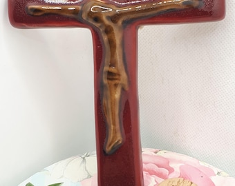 May include: A red ceramic crucifix with a brown Jesus figure. The crucifix is hanging on a white wall.