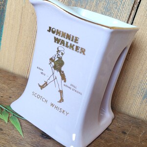Op de afbeelding: Een witte keramische karaf met een gouden rand en een Johnnie Walker logo. De karaf toont een man met een stok die loopt en de tekst "Born 1820 Still Going Strong Scotch Whisky".