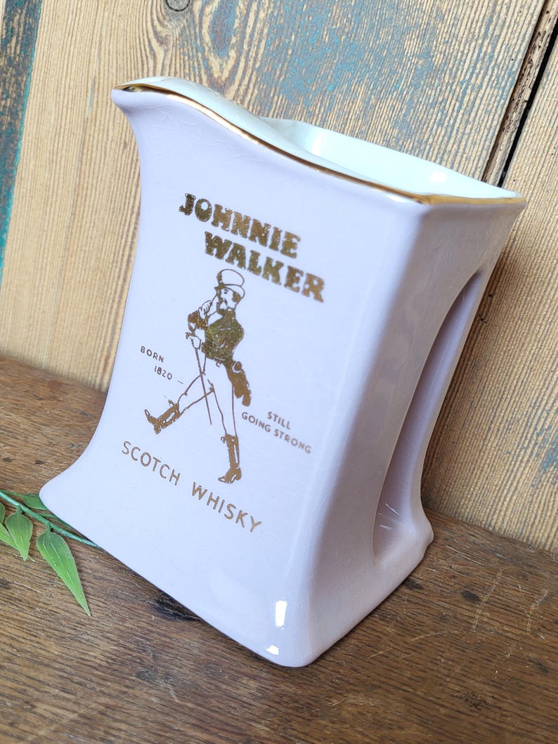 Op de afbeelding: Een witte keramische karaf met een gouden rand en een Johnnie Walker logo. De karaf toont een man met een stok die loopt en de tekst "Born 1820 Still Going Strong Scotch Whisky".