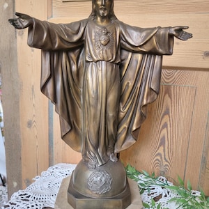 May include: A bronze-colored statue of Jesus with outstretched arms. The statue features detailed robes and a heart emblem. It stands on a globe-shaped base with a decorative emblem, mounted on a stone-like pedestal. The background includes wooden panels.