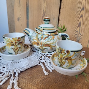 Puede incluir: Un juego de té de cerámica con una tetera y dos tazas con platillos. El juego tiene una base de color crema con un diseño floral en amarillo, marrón y verde. La tetera tiene una tapa y un asa a juego. Las tazas son pequeñas y redondas.