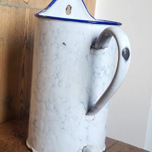 May include: Antique white enamel water jug with a blue rim and handle. The jug has a marbled pattern in blue and grey. It features a spout near the base and a curved handle. This vintage jug is likely for serving water or other drinks.
