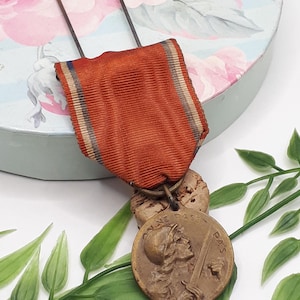 Op de afbeelding: Een vintage bronzen medaille met een rood lint. De medaille toont een profiel van een persoon die een zwaard vasthoudt, met de woorden "PAS" en "PAS" aan weerszijden. Het lint heeft blauwe, witte en beige strepen.