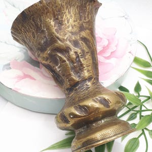 Puede incluir: Un jarrón de latón vintage con una superficie texturizada y una parte superior acampanada. El jarrón tiene una base bulbosa y una apariencia envejecida detallada. El jarrón es un artículo decorativo.