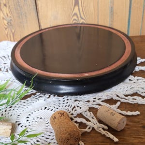 Peut inclure: Une base ronde en bois noir avec un rebord surélevé et un anneau intérieur brun foncé. La base est posée sur une surface en bois avec une dentelle crochetée blanche et deux bouchons de bouteille en liège.