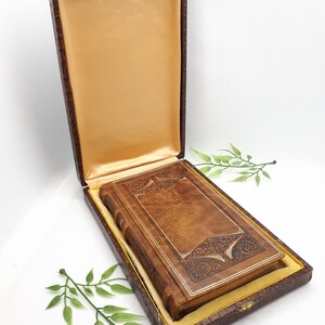 Puede incluir: Una caja de presentación rectangular de color marrón con tapa abatible y forro interior de satén dorado. Dentro de la caja descansa un libro rectangular marrón con intrincados patrones decorativos en relieve en la portada y el lomo.