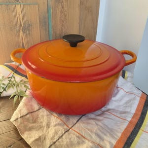 May include: A round enameled cast iron Dutch oven featuring a vibrant gradient finish that transitions from a deep red at the lid and base to a bright orange in the center. It includes two side handles and a black circular knob on the lid.