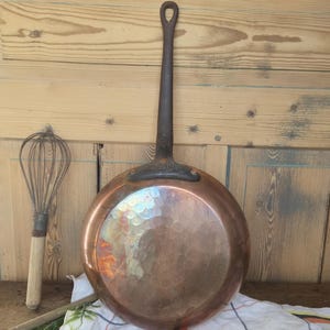 May include: A round, hammered copper pan with a long, dark, cast iron handle attached by two rivets. The pan features a polished, reflective surface with visible indentations from the hammering process and a circular hole at the handle end.