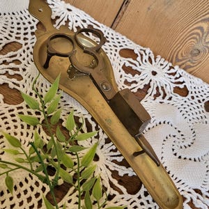 Puede incluir: Antiguo portavelas y tijeras de pared de latón. El portavelas tiene una forma rectangular larga y estrecha con un extremo redondeado. La parte superior del portavelas tiene un pequeño espacio redondo abierto para una vela. El portavelas también tiene un par de tijeras fijadas al lado.