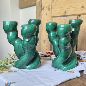 May include: A set of four green ceramic candle holders with a glossy finish. The candle holders have a unique, twisted design and are arranged on a white tablecloth with floral patterns. The background features wooden panels.