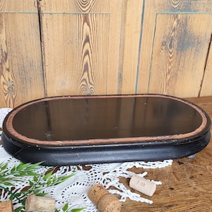 Peut inclure: Un présentoir en bois ovale noir avec un bord surélevé et une surface peinte en noir. Le présentoir a un aspect légèrement vieilli et est posé sur une surface en bois.