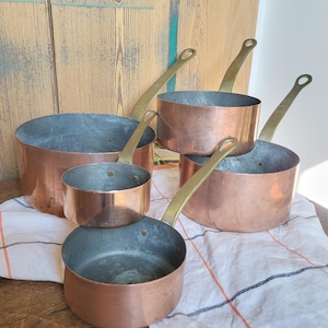 May include: A set of copper saucepans with brass handles. The pans are of varying sizes, with a large pot at the back and smaller ones in front. They are arranged on a white and orange striped cloth, set on a wooden surface.