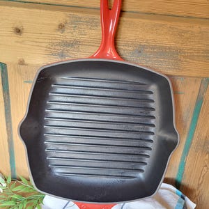 May include: A square, cast iron grill pan with a red handle. The pan has a black interior with raised grill lines. The handle is curved and has a hole for hanging. The pan is designed for grilling.
