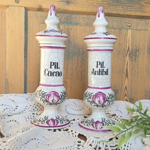May include: Two tall, off-white ceramic apothecary jars with a crackle glaze finish. Each jar features a pointed lid, a cylindrical body with black text reading Pil. Cacao and Pil. Antibil, and a base decorated with pink floral motifs and green leaves.
