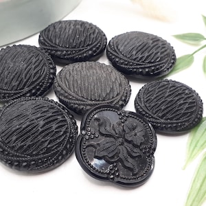Puede incluir: Una colección de siete botones negros redondos con patrones texturizados intrincados. Seis botones muestran un diseño de rejilla con bordes de cuentas, mientras que un botón presenta un motivo floral central con hojas y pequeñas bayas en relieve.