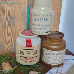 Puede incluir: Tres tarros de mostaza de cerámica vintage con etiquetas. Los tarros son marrones, beige y blancos. Las etiquetas están en francés e inglés. Los tarros están sobre una superficie de madera con una toalla a rayas blancas y naranjas.
