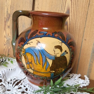 Może przedstawiać: Brązowy dzban ceramiczny z uchwytem, z ręcznie malowanym wzorem przedstawiającym osobę jeżdżącą konno. Wzór znajduje się w kółku z niebieskimi, żółtymi i zielonymi akcentami.