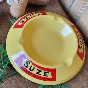 May include: Yellow metal ashtray with a red and black border and the text "Suze Liqueur du 4eme groupe" printed on the bottom.