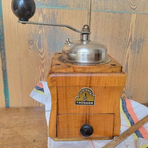 Puede incluir: Molinillo de café de madera vintage con tapa y manivela de metal. El molinillo tiene un cajón y un pomo negro. La marca "Armin Trösser" es visible. Una cuchara de madera y un paño estampado también están en la imagen.
