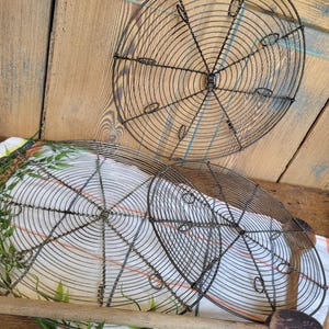 May include: Three round wire cooling racks, two are stacked on top of each other, and the third is on a wooden surface behind them. The racks are made of thin wire and have a circular design. The racks are a dark gray color and have a rustic look.