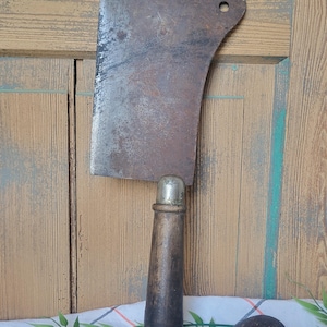 May include: A large, vintage meat cleaver with a dark, aged metal blade and a wooden handle. The blade has a hole near the top. The handle is dark brown with a silver metal cap. The cleaver rests on a white and red striped cloth.