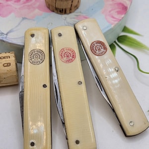 May include: Three vintage folding knives with cream-coloured handles. Each knife features a silver blade and a circular emblem with red text that reads "CANARD-DUCHESNE LUDES". The handles are secured with small, dark rivets.