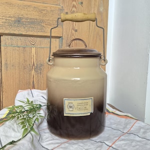 May include: A cylindrical enamel milk churn featuring a gradient colour scheme that transitions from a light beige top to a dark brown base. It includes a matching lid, a metal wire handle with a wooden grip, and a rectangular label with text.