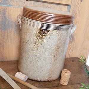 Op de afbeelding: Een grote, witte keramische pot met een bruin deksel en twee handvatten. De pot heeft een rustieke, verweerde uitstraling met bruine vlekken en strepen. De pot staat op een houten oppervlak met twee houten lepels en een kurk.