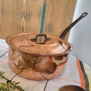 Op de afbeelding: Een koperen steelpan met een deksel en een lange handgreep. De pot heeft een glanzend, reflecterend oppervlak. Het deksel heeft een kleine handgreep. De handgreep is donker en gebogen. De pot staat op een wit en oranje gestreepte doek.