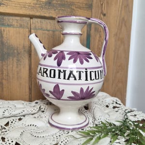 May include: A cream-colored ceramic apothecary pitcher featuring a rounded body, a narrow neck, a handle, and a spout. The vessel is decorated with hand-painted purple floral motifs and horizontal bands, with the word AROMATICUM in black lettering.
