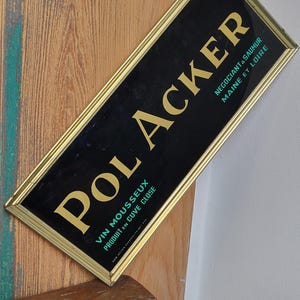 Peut inclure: Une enseigne dorée avec un fond noir et des lettres blanches qui disent "POLACKER VIN MOUSSEUX PRODUIT EN CUVÉ CLOSE" et "NÉGOCIANT À SAUMUR MAINE ET LOIRE".