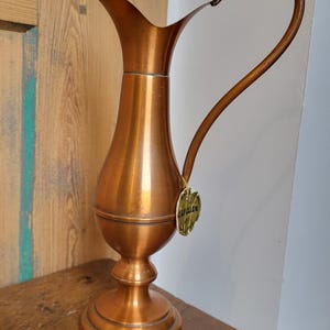 Könnte beinhalten: Eine kupferfarbene Kanne mit herzförmigem Griff. Die Kanne hat einen schlanken Hals und einen bauchigen Körper, der auf einem Sockel ruht. Ein kleines Etikett ist am Griff befestigt. Die Kanne steht auf einer Holzoberfläche.