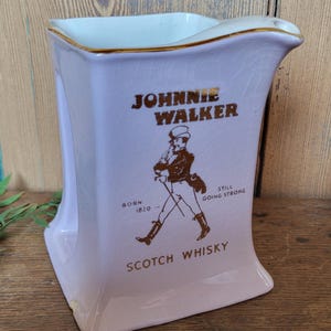 Op de afbeelding: Een paarse keramische kan met een gouden rand en het Johnnie Walker logo. Het logo toont een man in kilt die met een stok loopt en de tekst "Born 1820 - Still Going Strong" en "Scotch Whisky".