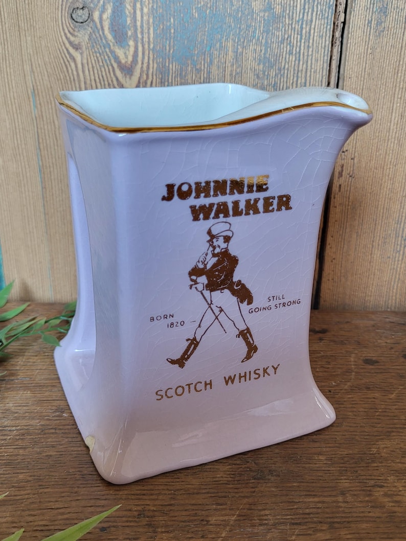 Op de afbeelding: Een paarse keramische kan met een gouden rand en het Johnnie Walker logo. Het logo toont een man in kilt die met een stok loopt en de tekst "Born 1820 - Still Going Strong" en "Scotch Whisky".
