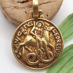 May include: A gold-colored pendant featuring a detailed relief of a figure on horseback slaying a serpent. The pendant has the inscription "SANCTVS GEORGIVS" around the edge. The pendant is attached to a small loop for a chain.
