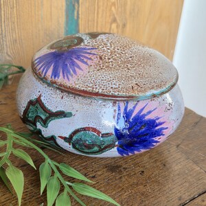 Könnte beinhalten: Ein Keramiktopf mit Deckel, mit einer gesprenkelten braunen und weißen Außenseite. Der Topf ist mit blauen und rosa Blumendesigns und grünen Akzenten verziert. Der Topf steht auf einer Holzoberfläche.