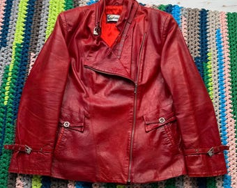 May include: A red leather jacket with a zip fastening and a silver buckle on the sleeve. The jacket has a collar and a "Cay-Cas" label on the front.