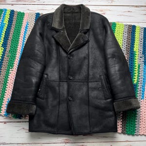 May include: A black leather coat with a shearling collar and cuffs. The coat has a button-up front and two pockets. The coat is displayed on a light wood surface with a colorful crocheted blanket in the background.