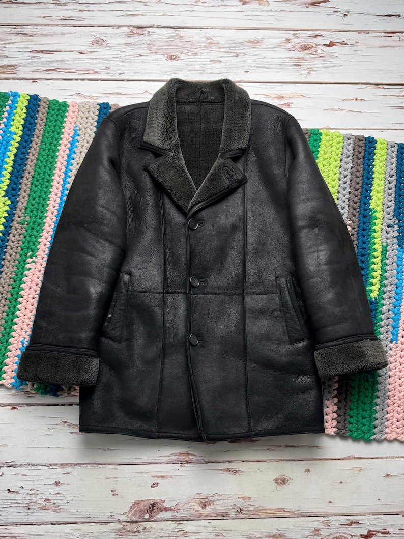 May include: A black leather coat with a shearling collar and cuffs. The coat has a button-up front and two pockets. The coat is displayed on a light wood surface with a colorful crocheted blanket in the background.