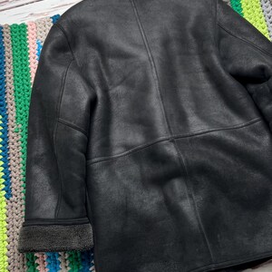 May include: A black jacket with a textured matte finish, featuring a fold-over collar and cuffs lined with dark gray fleece. The garment has visible vertical and horizontal seam detailing across the back and sleeves, providing a structured look.