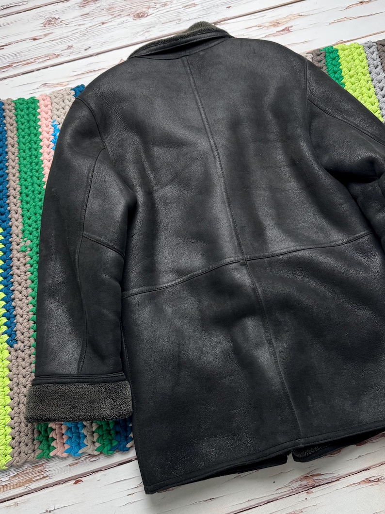 May include: A black jacket with a textured matte finish, featuring a fold-over collar and cuffs lined with dark gray fleece. The garment has visible vertical and horizontal seam detailing across the back and sleeves, providing a structured look.