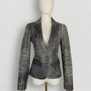 May include: A charcoal gray blazer with a distressed, weathered finish, featuring a single-button closure, notched lapels, and two rectangular flap pockets at the waist. The garment is displayed on a mannequin, showcasing long sleeves and a tailored fit.