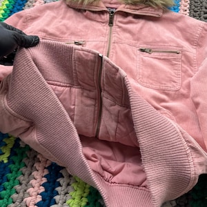 May include: A pink corduroy jacket with a faux fur hood. The jacket has a zipper closure and two pockets.