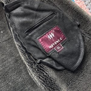 May include: Close-up of a black leather jacket pocket with a zipper. The pocket has a label that reads "MIYINUO" and "XL". The jacket has a dark gray, textured, fuzzy lining.