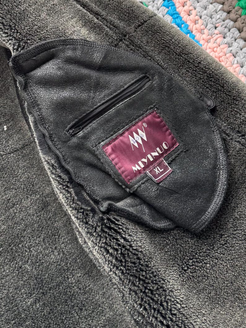 May include: Close-up of a black leather jacket pocket with a zipper. The pocket has a label that reads "MIYINUO" and "XL". The jacket has a dark gray, textured, fuzzy lining.