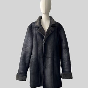 May include: A black shearling coat with a gray shearling collar and cuffs. The coat has a button-up front and is displayed on a mannequin. The coat is a classic style, suitable for cold weather.